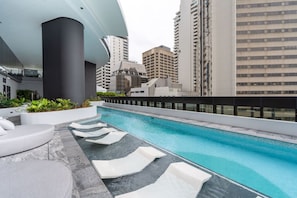 Un espace piscine aménagé sur le toit, avec des chaises longues blanches, une structure architecturale moderne et des immeubles de grande hauteur qui l’entourent.