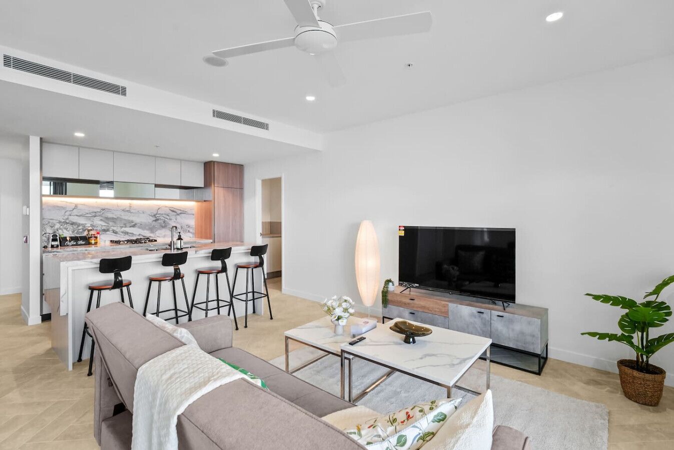A modern living room with a sofa, a coffee table, and a TV. There is a kitchen area with a bar and stools.