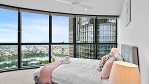 A bedroom with a large bed, a ceiling fan, and a view of a cityscape through large windows.
