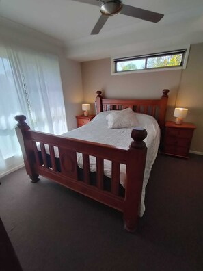 A bedroom with a wooden bed, two bedside tables with lamps, a ceiling fan, and a window with a view of greenery. - Cosy, Modern, 4BR Home In Mullumbimby (Mullumbimby)