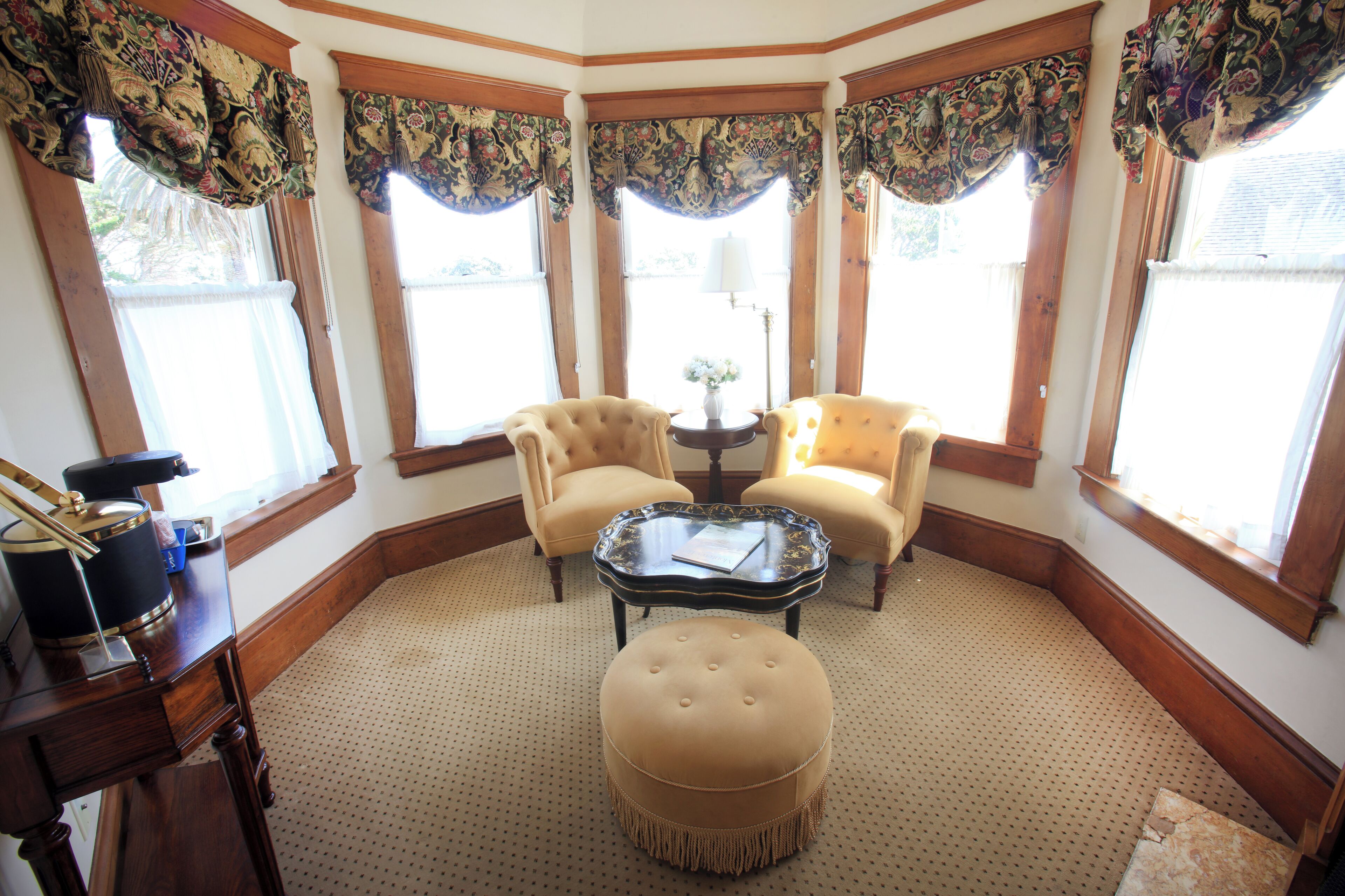 A corner room with two armchairs, a round ottoman, a small table, and a piano.