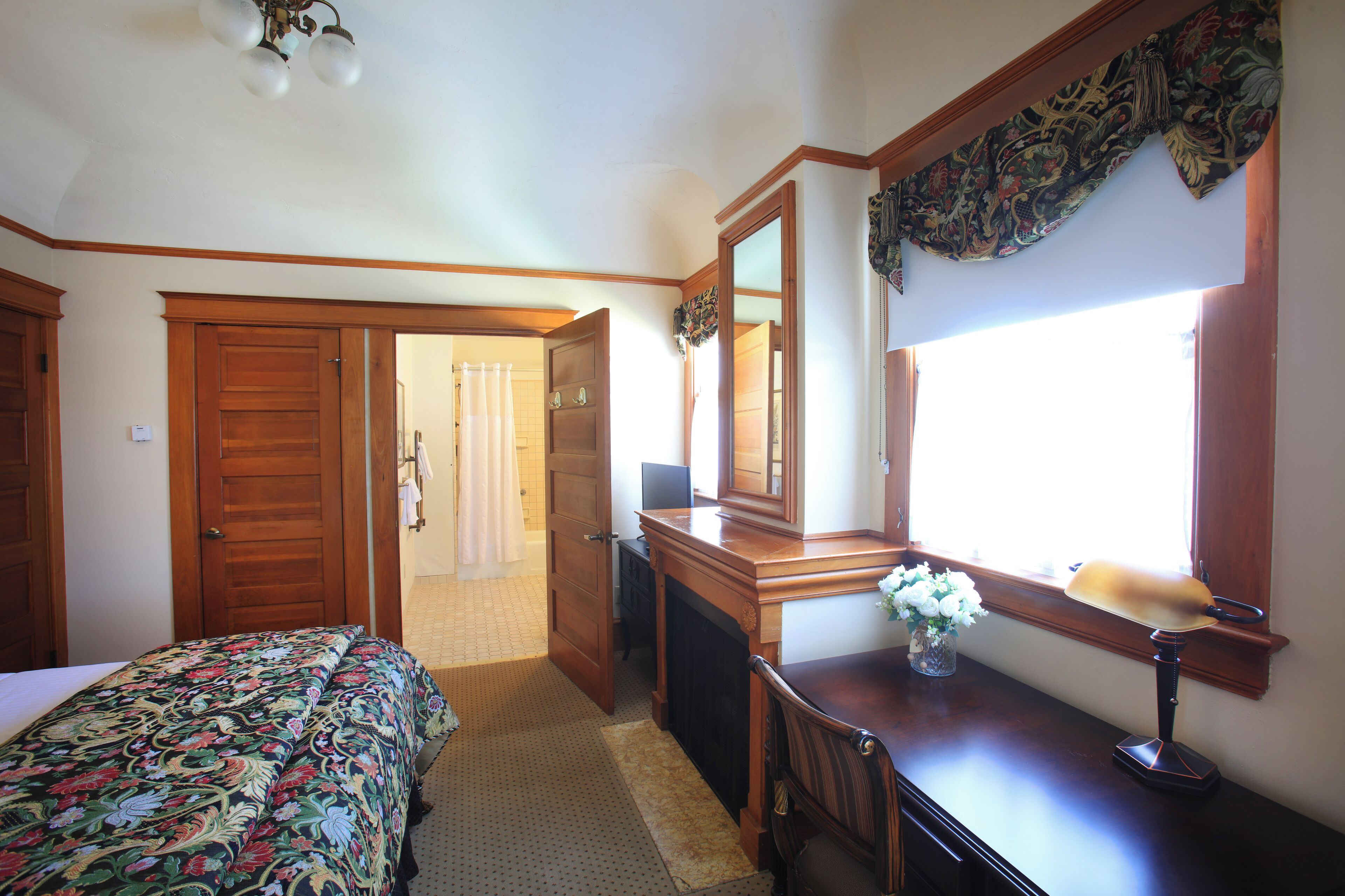 Un dormitorio con cama, escritorio con lámpara, espejo y ventana con cortinas.
