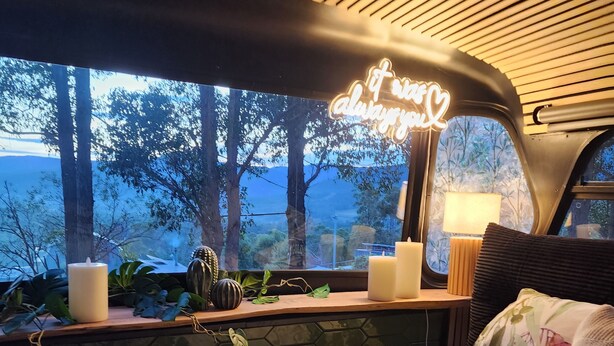 A cozy interior with a bed, a window with a neon sign, and a view of trees outside. - Unique, Exciting Bus Located on 63 acres with a Cosy woodheater. Luxury. (Mountain River)