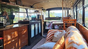A cozy interior with a curved ceiling, a sofa with patterned cushions, a wooden cabinet, and a window offering a view of the outdoors. - Unique, Exciting Bus Located on 63 acres with a Cosy woodheater. Luxury. (Mountain River)