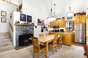 A kitchen with a stone fireplace, wooden dining table, and chairs. There are American flags and framed pictures on the walls. - 27 Circle Four Cabin - AC & 8 SHARC Passes! (Sunriver)