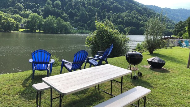 A picnic setup with a table and chairs by a lake, surrounded by lush greenery and hills.
