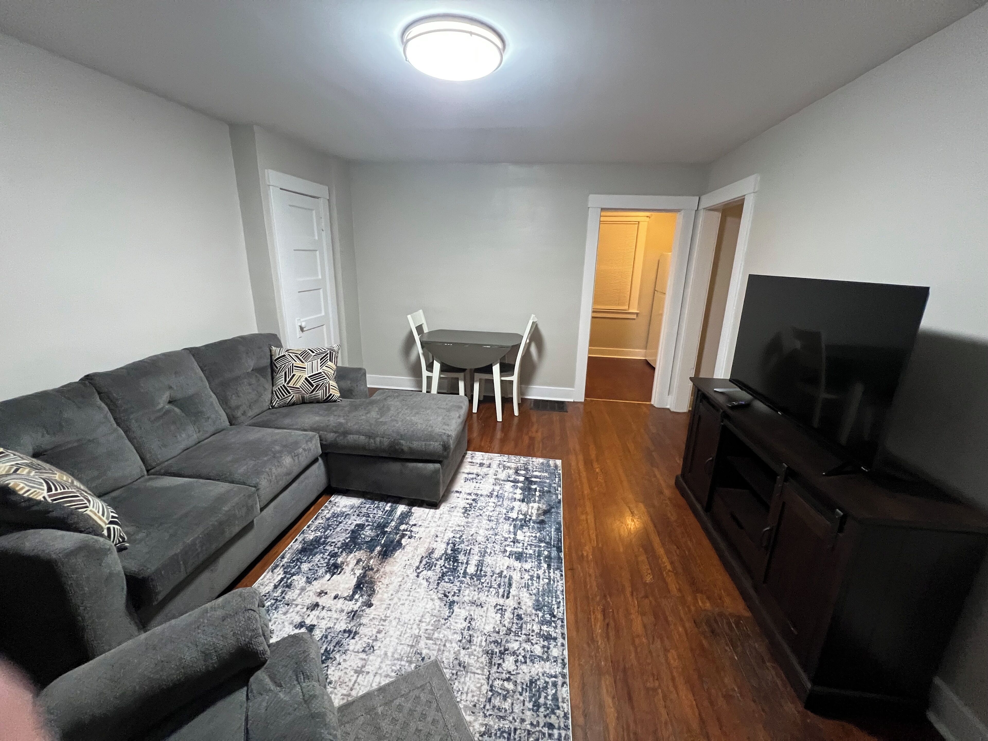 A living room with a sectional sofa, a flat-screen TV, a dining table with chairs, and a hallway leading to another room.