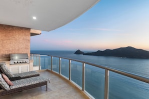 A balcony with a grill, lounge chairs, and a view of the sea.