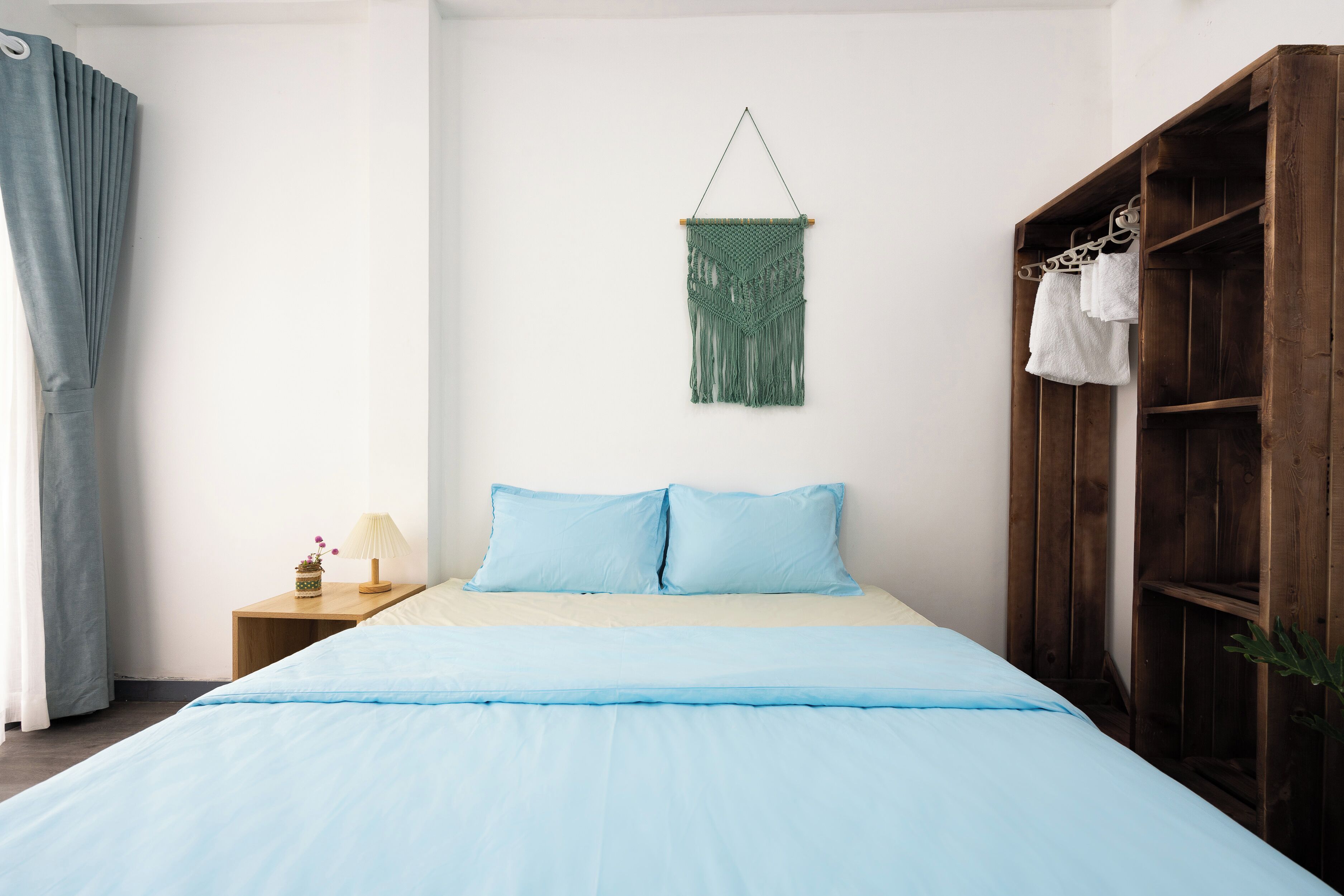 Un dormitorio con cama, armario de madera, mesita de noche, lámpara y un tapiz de macramé en la pared.