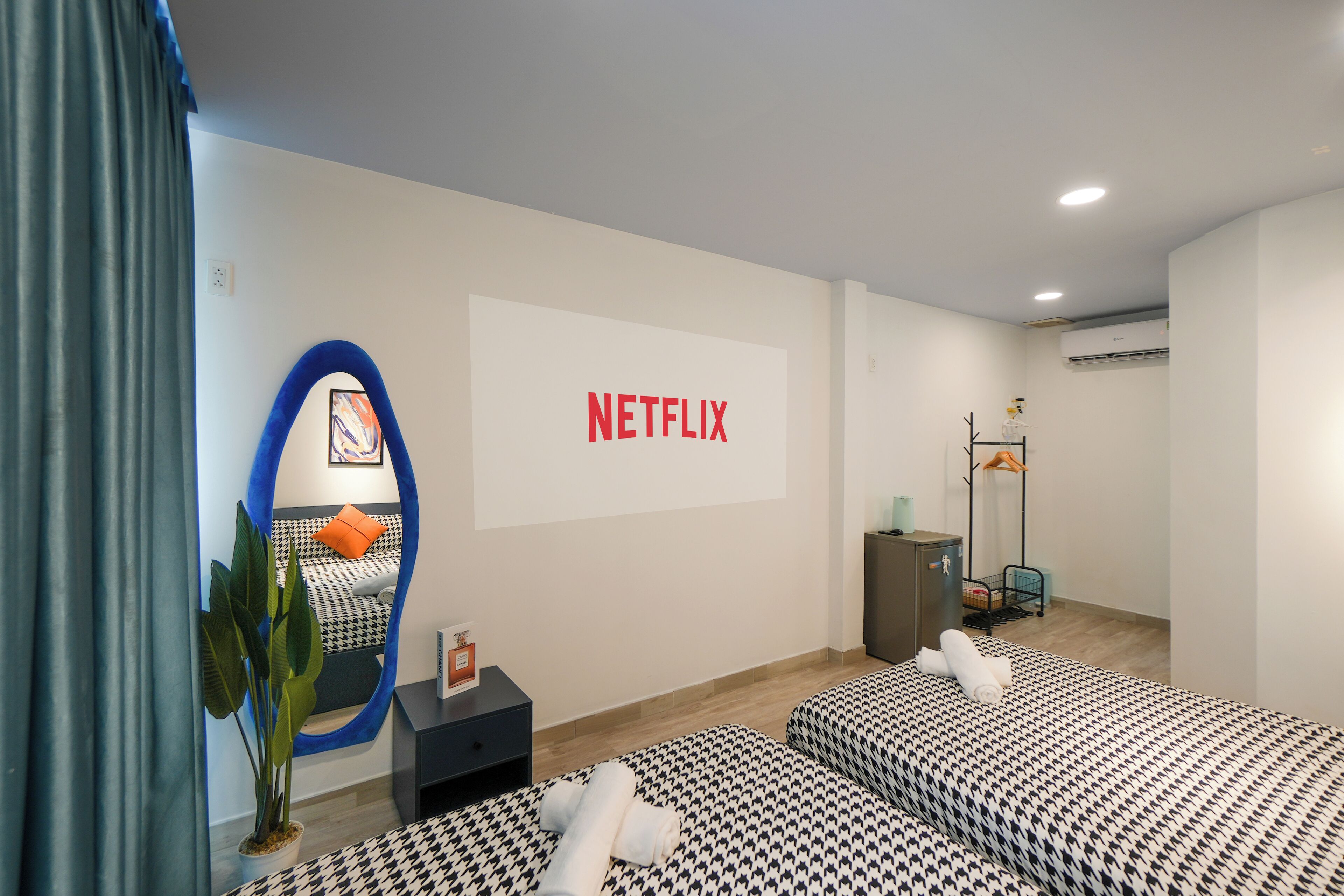 A hotel room with two beds, a large mirror, a wall-mounted TV, a plant, and a small table with a bottle.