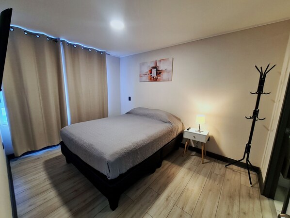 A bedroom with a bed, a nightstand, a lamp, a coat rack, and a painting on the wall.