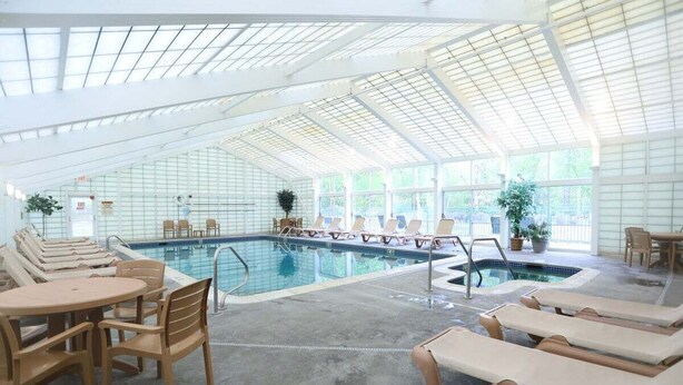 An indoor swimming pool with lounge chairs and tables, surrounded by glass walls and a high ceiling. - Rivergreen 301, Lincoln (Lincoln)