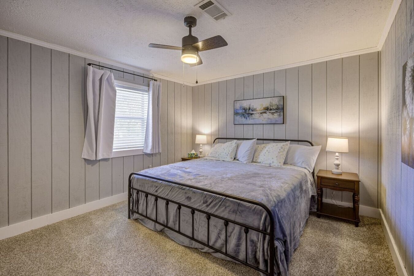 A bedroom with a bed, a ceiling fan, a window with curtains, a bedside table with a lamp, and a painting on the wall.