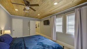 Un dormitorio con cama, ventilador de techo, una puerta, ventana con cortinas y televisor montado en la pared.