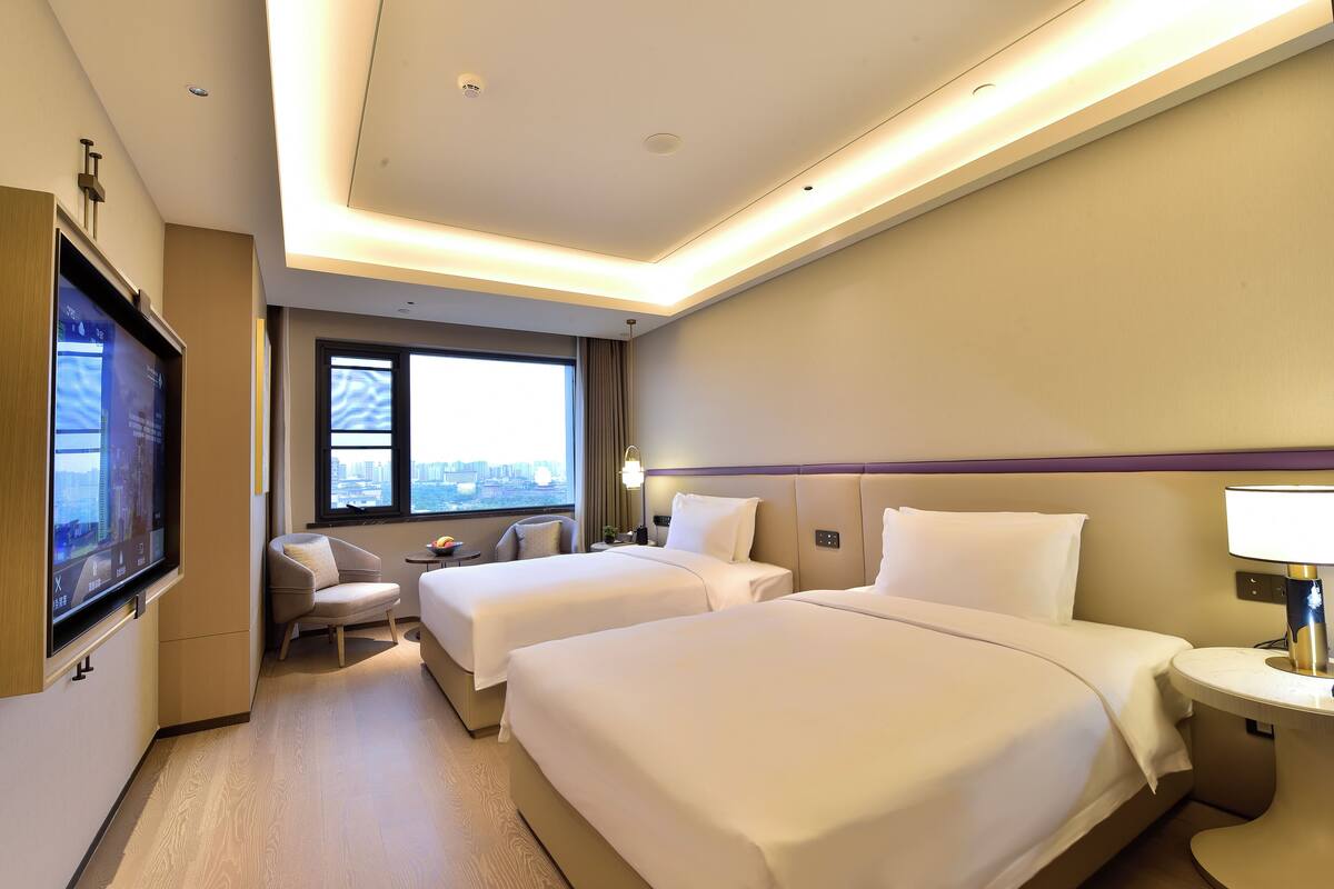 A hotel room with two beds, a TV, a desk, and a window with a city view.