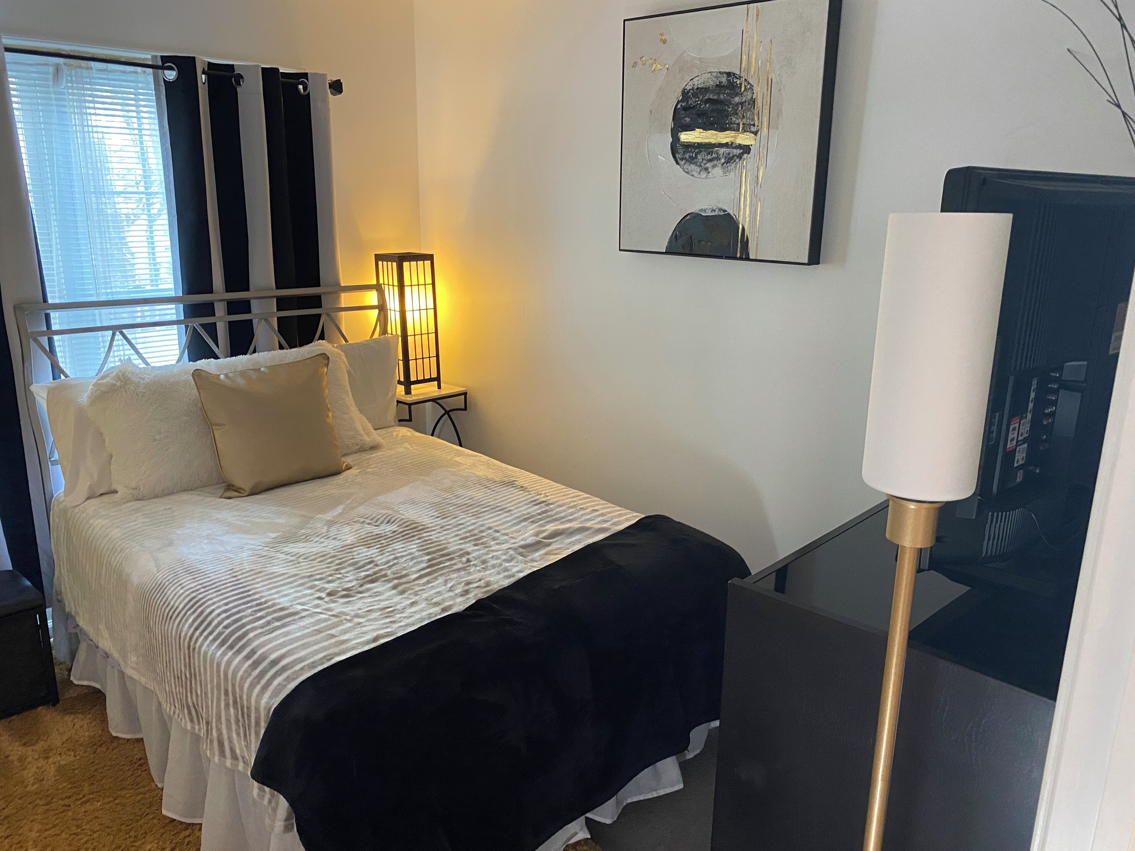 Ein ordentlich bezogenes Bett mit weißer und schwarzer Bettwäsche, eine Nachttischlampe und ein gerahmtes abstraktes Kunstwerk an der Wand.
