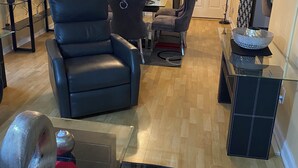 Una sala de estar con un sofá de cuero negro, una mesa de centro de vidrio y un comedor con una araña.