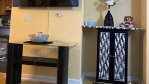 Una sala de estar con un televisor de pantalla plana montado en la pared, una mesa de centro de vidrio con un cuenco decorativo, un mueble con estampado y un oso de peluche, y un florero con flores.