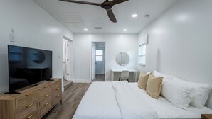 A modern bedroom with a large bed, a TV on a wooden dresser, a ceiling fan, and a desk with a chair.