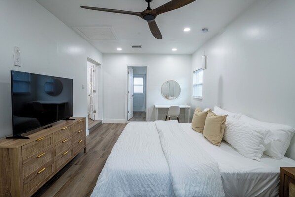 Un dormitorio moderno con una cama grande, un televisor sobre un aparador de madera, un ventilador de techo y un escritorio con una silla.