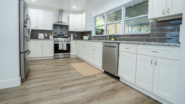 A modern kitchen with white cabinets, a stainless steel refrigerator, a dishwasher, and a large window.
