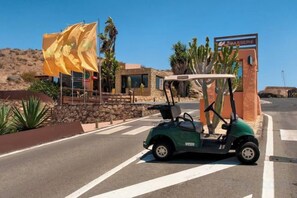 A golf cart parked in a parking lot with a building and a large yellow flag in the background. - Vista Golf 30 Salobre by VillaGranCanaria (Comarca Sur)