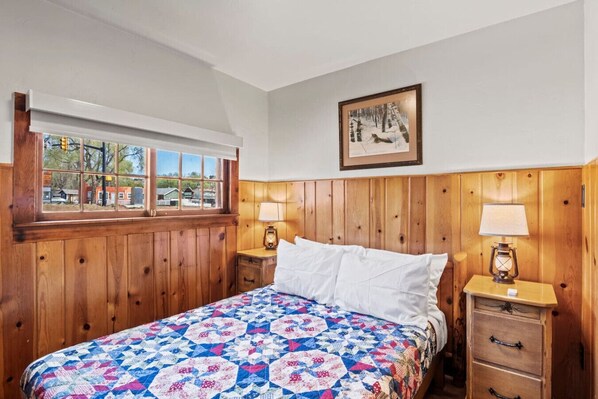 A bedroom with wooden paneling, a bed with a colorful quilt, two bedside tables with lamps, a window with a view, and a framed picture on the wall. - Timber Lodge Cabin #1 (Colorado Springs)