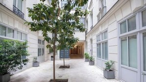 A narrow corridor with potted plants and a central tree, flanked by buildings with large windows and a door at the end. - Champs Elysees Charming (2 Bedrooms and 1 Living Room) (Paris)