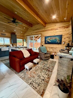 A cozy cabin interior with a red couch, a grey armchair, a kitchen area, and a large wooden ceiling.
