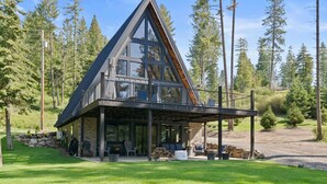A modern two-story cabin with a large glass balcony, surrounded by a lush green landscape and tall trees. - Lake Views At Rockford Bay - The CDA-frame (Coeur d`Alene)