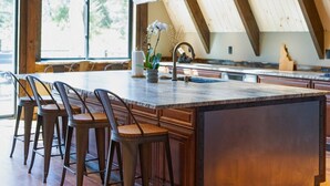 A modern kitchen with a large island, wooden bar stools, and a view of the outdoors through large windows. - Lake Views At Rockford Bay - The CDA-frame (Coeur d`Alene)