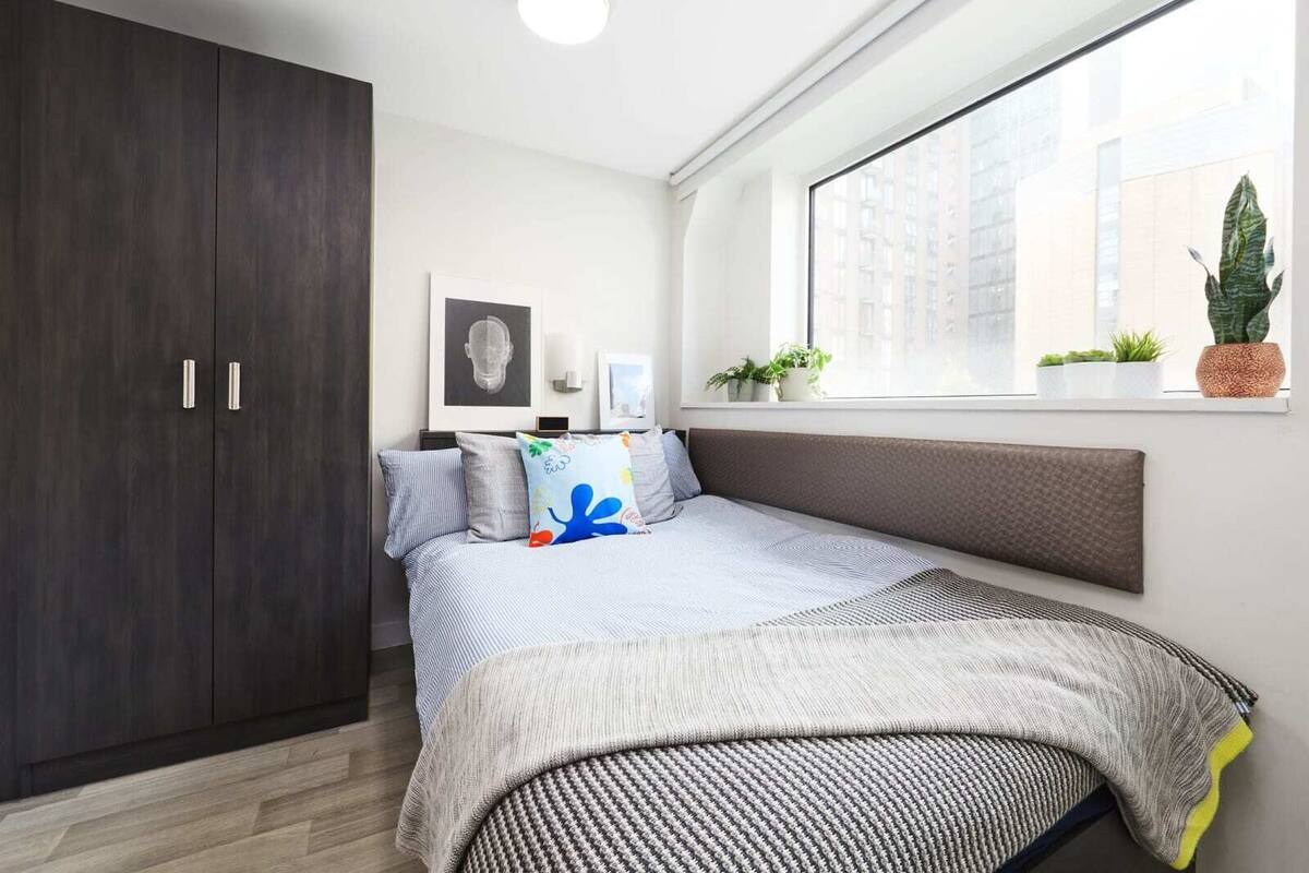 Ein Schlafzimmer mit großem Kleiderschrank, einem Bett mit gemustertem Überwurf und einem Fenster mit Pflanzen, das auf die Stadt blickt.