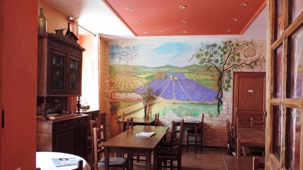A dining area with a mural, wooden furniture, and a kitchen area with cabinets. - Hotel Provence by Adonis (Digne-les-Bains)