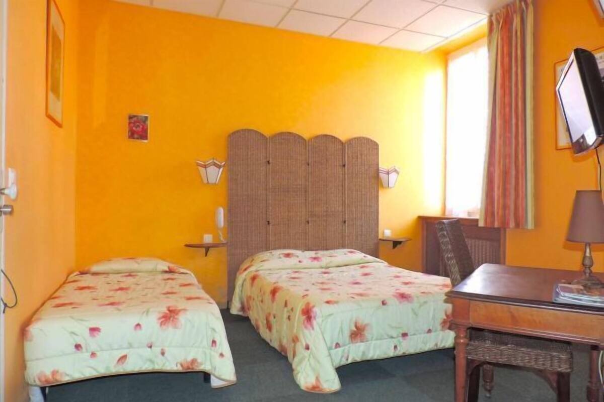Quarto com duas camas, uma escrivaninha, uma cadeira, uma televisão e uma janela com cortinas.