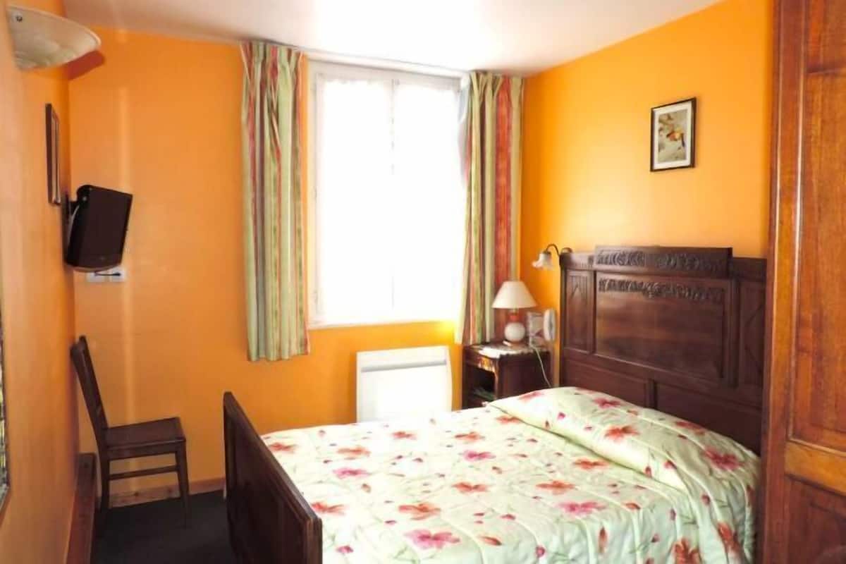 Quarto com uma cama, uma cadeira, uma escrivaninha com luminária, televisão e janela com cortinas.