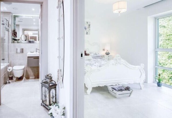 Una camera da letto con un letto bianco decorato, un comodino con una lampada e vista attraverso un'ampia finestra.