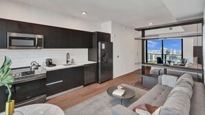 A modern kitchen with dark cabinets, a stainless steel microwave, and a refrigerator. There is a dining area with a sofa and a coffee table. - Luxury Miami Studio Bay Views | 37th Fl (Miami)