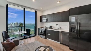 A modern kitchen with dark cabinets, a stainless steel refrigerator, and a large window offering a city view. - Luxury Miami Studio Bay Views | 37th Fl (Miami)