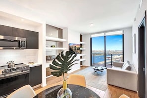 A modern kitchen with a built-in microwave and stove, a dining area with a table and chairs, and a living room with a sofa and a large window offering a view.