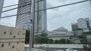 A view through a window with a metal grid, featuring a tall building with a curved facade, a parking lot with several cars, and a clear sky. - 15min Shinjuku/5min Hatsudai(OperaCityDirect).Quiet,perfect place/Early check-in (Shinjuku City)