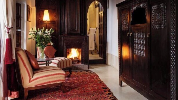 A cozy room with a fireplace, a striped armchair, a red rug, and a wooden cabinet. - Riad Azzar (Marrakech)