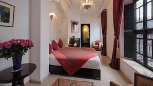A hotel room with a bed, a small table with a vase of flowers, a window with curtains, and a view of the outside. - Riad Azzar (Marrakech)