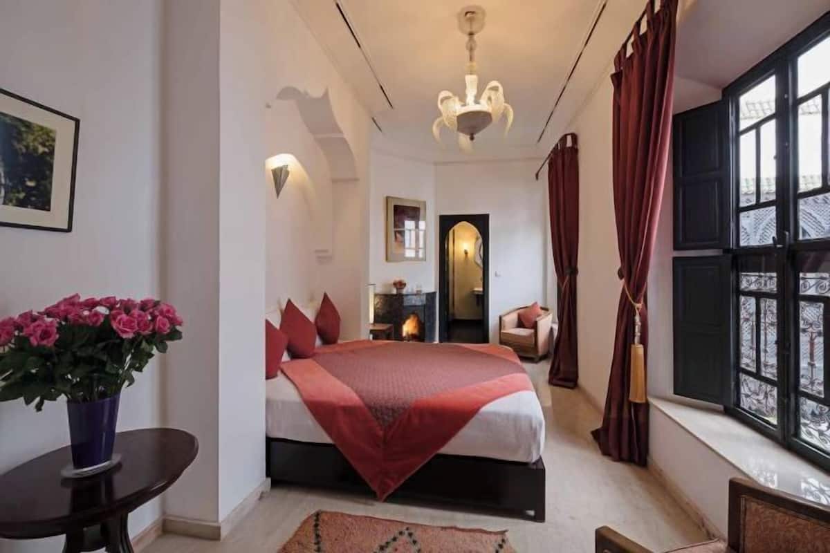 Ein Hotelzimmer mit Bett, einem kleinen Tisch mit Blumenvase, einem Fenster mit Vorhängen und Blick ins Freie.