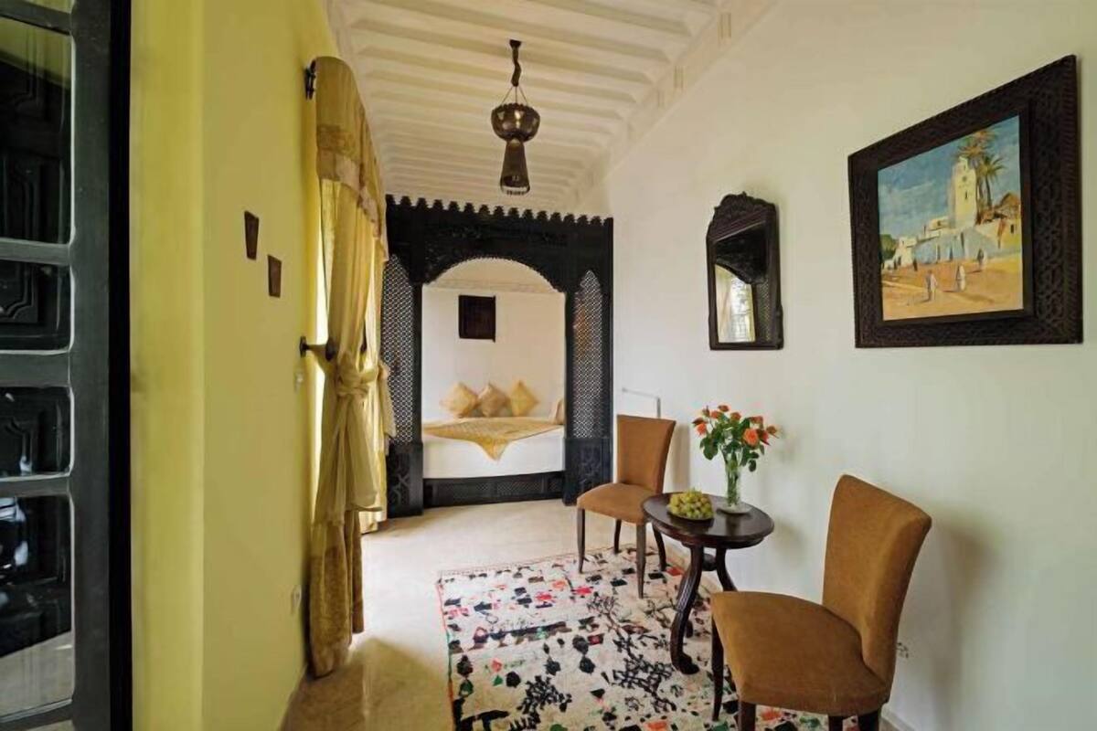 Ein Zimmer mit einem Bett, einem Sessel, einem kleinen Tisch mit Blumen und einem Bild an der Wand.