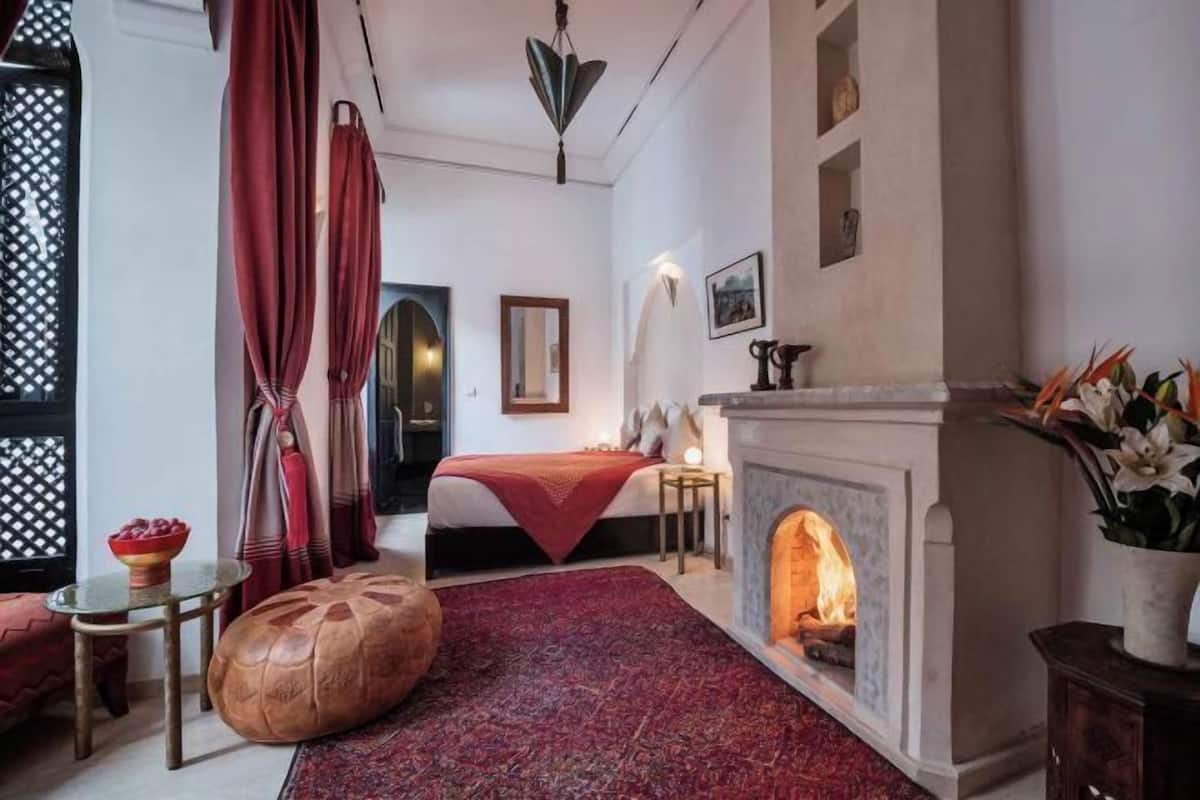 Ein Zimmer mit einem Bett, einem Kamin, einem runden Ottomanen, einem kleinen Tisch und einem dekorativen Spiegel.