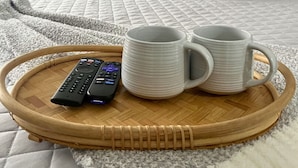 A neatly made bed with a green headboard, two pillows, and a white blanket. A tray with two white mugs and a remote control is placed on the bed.