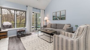 A living room with a sofa, armchair, coffee table, and a view of the outdoors through large windows.
