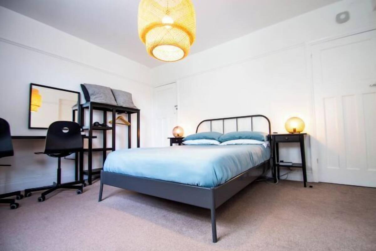 A bedroom with a bed, a desk, a chair, a hanging light fixture, and a door.