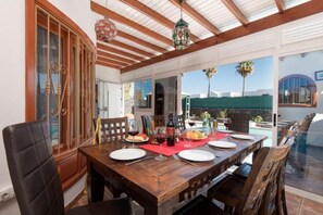 Un coin repas avec une table en bois dressée pour quatre personnes, une porte vitrée donnant sur l’extérieur et une vue sur des palmiers.