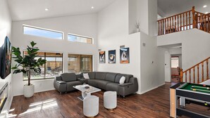 A spacious living room with a grey sectional sofa, a pool table, and a large window overlooking a cityscape.